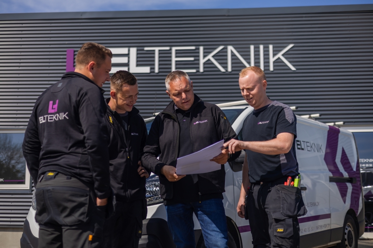 Fire elektrikere står foran en firmabil og gennemgår tegninger sammen udenfor virksomhedens bygning. Billedet viser samarbejde og planlægning af en opgave før arbejdet går i gang.