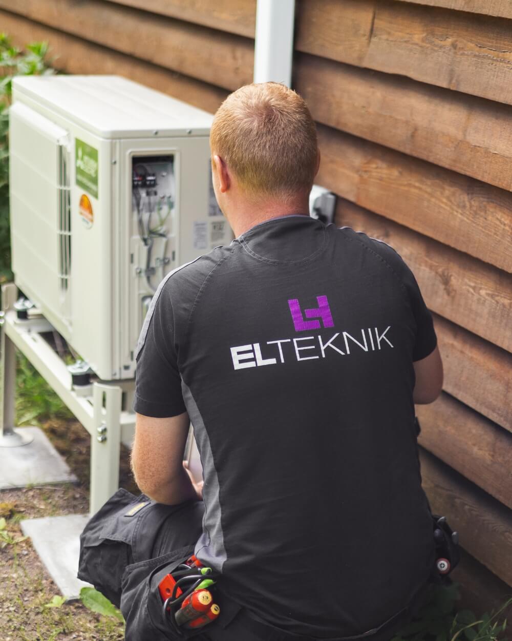 Elektriker fra ELTEKNIK sidder ved en udendørs varmepumpe og arbejder på installationen ved siden af en træbeklædt husmur. Situationen viser service eller montering af varmepumpeanlæg ved en bolig.